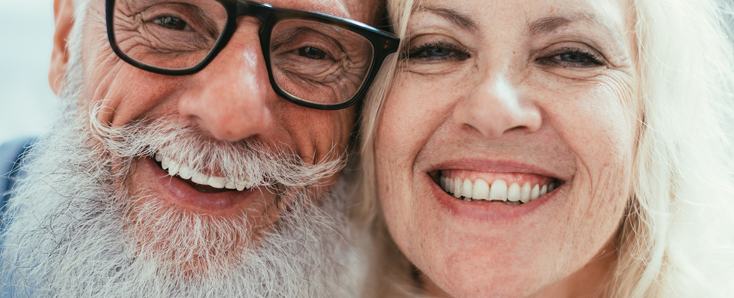 older couple of a man with dark-rimmed eyeglasses, a mustache, and a beard next to a woman with light hair smiling
