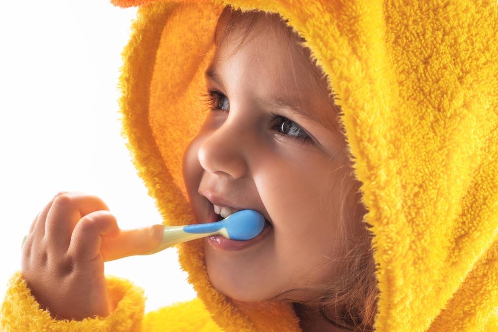 blonde-haired child wearing a yellow fuzzy hoodie brushing her teeth with a blue and yellow toothbrush and smiling